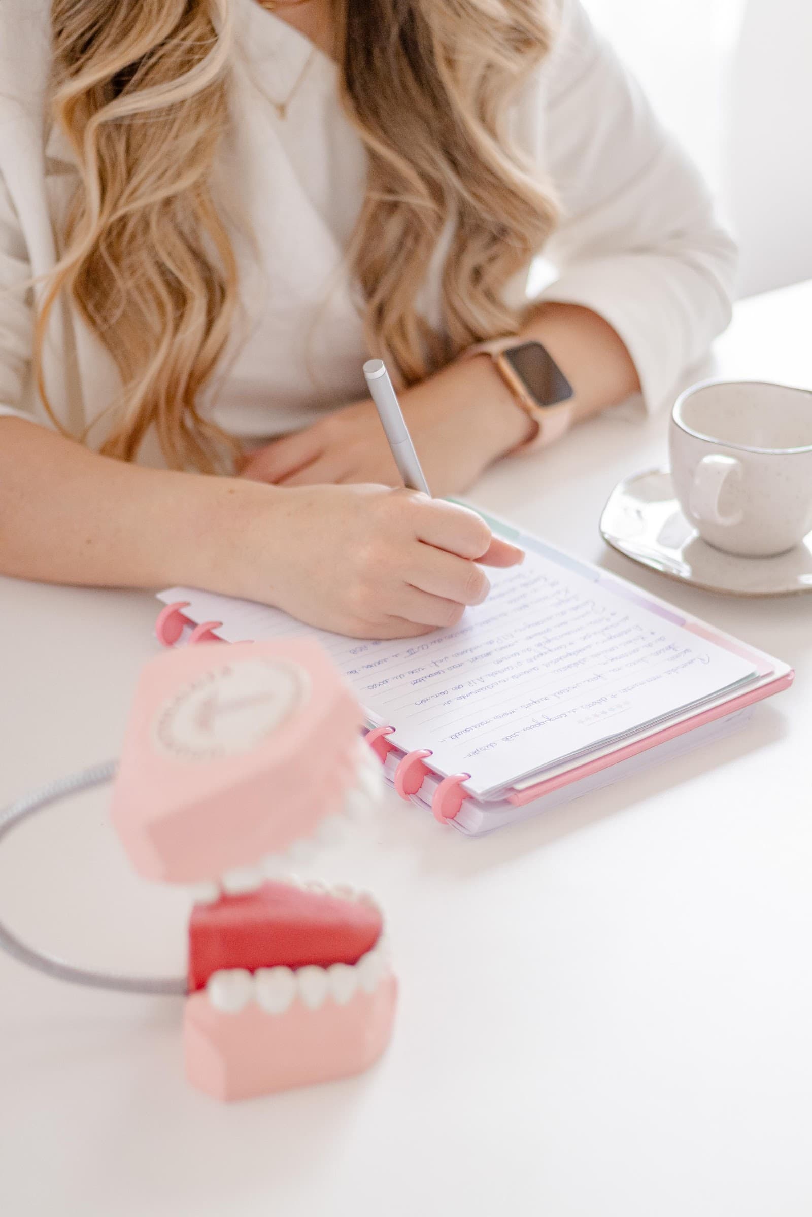 Mão da dentista anotando num caderno rosa, com um modelo de arcada dentária na mesa